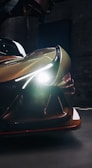 A close-up view of a sleek, futuristic car with its headlights on, illuminating the dark surroundings. The car's design features sharp lines and glossy surfaces, emphasizing its modern and dynamic appearance.