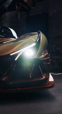 A close-up view of a sleek, futuristic car with its headlights on, illuminating the dark surroundings. The car's design features sharp lines and glossy surfaces, emphasizing its modern and dynamic appearance.