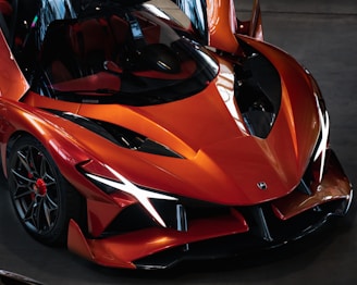 A sleek, aerodynamic sports car with sharp, angular lines and a vibrant orange color. The design includes prominent front fenders, a low-slung body, and distinctive black accents. The vehicle features advanced wheel rims and glossy reflections on the surface, highlighting its modern and luxurious appearance.