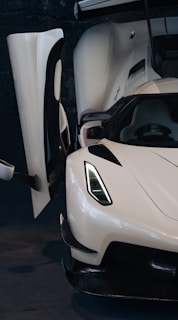 A sleek white sports car with an open gullwing door, showcasing an aerodynamic design. The car's angular headlights and smooth curves emphasize its modern and high-performance features.