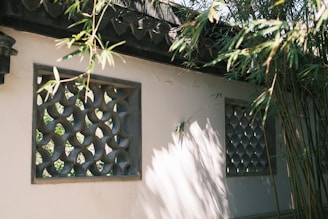 Sunlight filters through bamboo leaves, casting shadows on a cream-colored wall with two carved openings. The lattice patterns in the windows are ornate, providing a glimpse of greenery beyond. The tranquil setting is enhanced by the play of light and shadow on the textured surface.