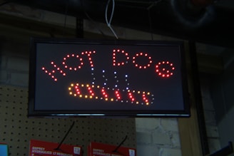 An LED sign displaying the words 'HOT DOG' in red lights with decorative elements represented by small dots. The sign is mounted on a background of pegboard and brick wall.
