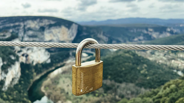 A brass padlock is secured to a metal cable overlooking a scenic landscape with rolling hills, a river, and cloudy skies in the background.