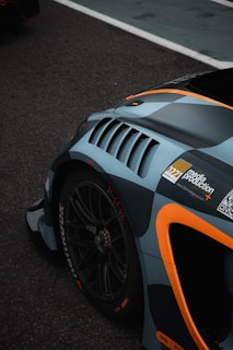 A close-up of a sports car's rear section with aerodynamic features. The car is adorned with racing decals and has a sleek, modern design. The tire's branding is visible, and a QR code is present on the car's body.