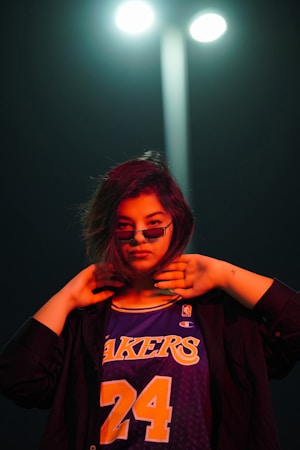 A person stands confidently under a streetlight, wearing a Los Angeles Lakers basketball jersey with the number 24. The lighting casts a dramatic effect with red and blue tones. The individual has short hair and is wearing sunglasses, with a stylish pose and hands near the face.