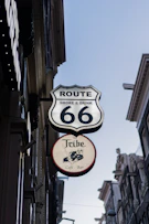 A vertical street view featuring a prominent sign for Route 66, labeled 'Smoke & Drink' in bold letters. Below it, there is another sign for 'The Tribe Cafe - Bar' with an illustration of a person holding a spear. The signs are attached to buildings lining a narrow street.
