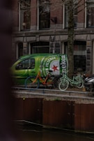 A green van with a large Heineken logo is parked on a street beside a canal. Two bicycles, one orange with blue wheels and one mint green, are leaning against the van. The background features a traditional brick building with multiple windows.