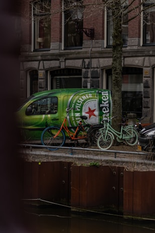 A green van with a large Heineken logo is parked on a street beside a canal. Two bicycles, one orange with blue wheels and one mint green, are leaning against the van. The background features a traditional brick building with multiple windows.