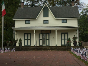 A two-story house with a symmetrical design, featuring a dark gray roof and beige siding. It is surrounded by a paved area with numerous small American flags lined up in rows. There are flower pots and bushes in the vicinity, and a flagpole with an Italian flag can be seen on the left side. The setting is likely during fall as indicated by the leaves on the ground and the trees in the background.