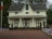 A two-story house with a symmetrical design, featuring a dark gray roof and beige siding. It is surrounded by a paved area with numerous small American flags lined up in rows. There are flower pots and bushes in the vicinity, and a flagpole with an Italian flag can be seen on the left side. The setting is likely during fall as indicated by the leaves on the ground and the trees in the background.