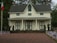 A two-story house with a symmetrical design, featuring a dark gray roof and beige siding. It is surrounded by a paved area with numerous small American flags lined up in rows. There are flower pots and bushes in the vicinity, and a flagpole with an Italian flag can be seen on the left side. The setting is likely during fall as indicated by the leaves on the ground and the trees in the background.