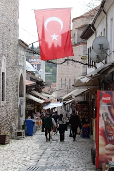 A narrow cobblestone street flanked by historic stone buildings, with people walking along the path. A large Turkish flag hangs overhead, and various shops line the street. A Coca-Cola advertisement is visible on the right, alongside goods and market offerings. The atmosphere suggests a blend of traditional and modern elements.