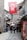 A narrow cobblestone street flanked by historic stone buildings, with people walking along the path. A large Turkish flag hangs overhead, and various shops line the street. A Coca-Cola advertisement is visible on the right, alongside goods and market offerings. The atmosphere suggests a blend of traditional and modern elements.
