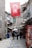 A narrow cobblestone street flanked by historic stone buildings, with people walking along the path. A large Turkish flag hangs overhead, and various shops line the street. A Coca-Cola advertisement is visible on the right, alongside goods and market offerings. The atmosphere suggests a blend of traditional and modern elements.