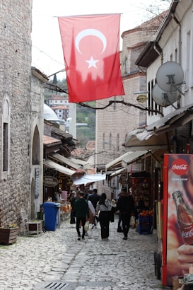 A narrow cobblestone street flanked by historic stone buildings, with people walking along the path. A large Turkish flag hangs overhead, and various shops line the street. A Coca-Cola advertisement is visible on the right, alongside goods and market offerings. The atmosphere suggests a blend of traditional and modern elements.
