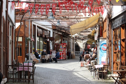 A narrow, cobblestone street lined with wooden storefronts and outdoor seating. Turkish flags hang overhead, adding a festive and cultural touch. People are seated at tables, engaging in conversation or relaxing, creating a lively yet cozy atmosphere.