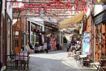 A narrow, cobblestone street lined with wooden storefronts and outdoor seating. Turkish flags hang overhead, adding a festive and cultural touch. People are seated at tables, engaging in conversation or relaxing, creating a lively yet cozy atmosphere.