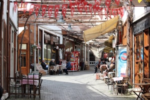 A narrow, cobblestone street lined with wooden storefronts and outdoor seating. Turkish flags hang overhead, adding a festive and cultural touch. People are seated at tables, engaging in conversation or relaxing, creating a lively yet cozy atmosphere.