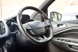 The interior of a car showing the steering wheel with a Ford emblem, dashboard, and driver's side door. The steering wheel has a textured grip with red stitching and multiple controls. The car door features a handle and control buttons for windows.