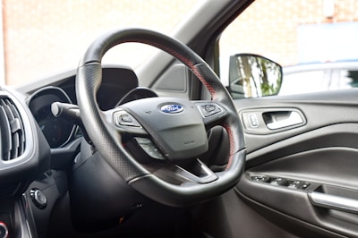 The interior of a car showing the steering wheel with a Ford emblem, dashboard, and driver's side door. The steering wheel has a textured grip with red stitching and multiple controls. The car door features a handle and control buttons for windows.
