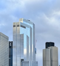 Tall modern skyscrapers with reflective glass facades stand against a cloudy sky. The architecture features geometric designs with vertical lines and sharp angles. A construction crane is visible in the background.