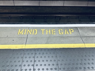 A train platform edge with a warning message painted on the ground in yellow that reads 'Mind the Gap'. The area consists of a textured surface with dotted patterns and a distinct yellow line indicating safety boundaries.
