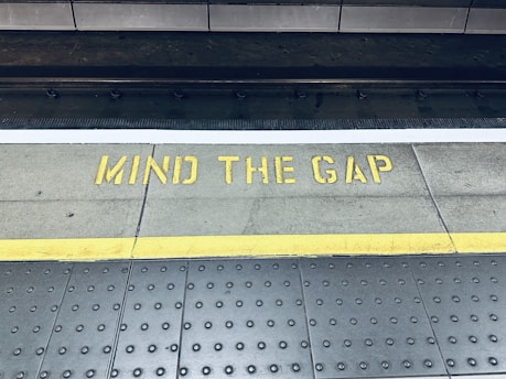 A train platform edge with a warning message painted on the ground in yellow that reads 'Mind the Gap'. The area consists of a textured surface with dotted patterns and a distinct yellow line indicating safety boundaries.