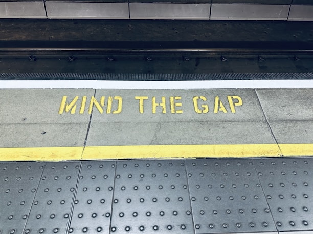 A train platform edge with a warning message painted on the ground in yellow that reads 'Mind the Gap'. The area consists of a textured surface with dotted patterns and a distinct yellow line indicating safety boundaries.