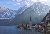 A picturesque lakeside village rests at the foot of towering, snow-capped mountains. The architecture is characterized by a prominent church steeple, surrounded by charming, traditional houses with sloping roofs. The calm lake reflects the buildings and the mountain scenery, adding to the serene atmosphere.