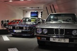 Several classic BMW cars are on display in a showroom. The cars are positioned side by side, showcasing their vintage design. A red car and an engine are visible in the background along with several colorful car panels mounted on the wall.