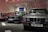 Several classic BMW cars are on display in a showroom. The cars are positioned side by side, showcasing their vintage design. A red car and an engine are visible in the background along with several colorful car panels mounted on the wall.
