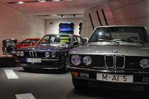 Several classic BMW cars are on display in a showroom. The cars are positioned side by side, showcasing their vintage design. A red car and an engine are visible in the background along with several colorful car panels mounted on the wall.