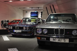 Several classic BMW cars are on display in a showroom. The cars are positioned side by side, showcasing their vintage design. A red car and an engine are visible in the background along with several colorful car panels mounted on the wall.