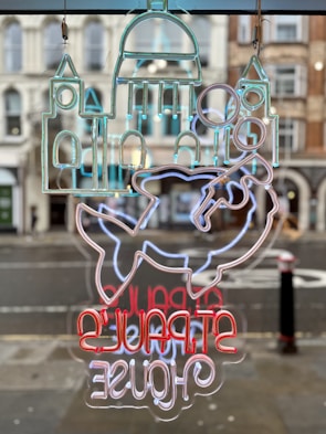 The image features a neon sign with an abstract design depicting elements like a building structure and a fish. The sign includes text that is mirrored, displayed in vibrant colors. It hangs against a backdrop of a street view with classic architectural buildings in the background.