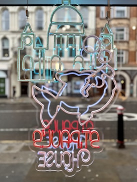 The image features a neon sign with an abstract design depicting elements like a building structure and a fish. The sign includes text that is mirrored, displayed in vibrant colors. It hangs against a backdrop of a street view with classic architectural buildings in the background.