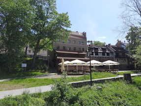 A picturesque outdoor café area with tables and large sun umbrellas situated in front of a series of old-style buildings. A well-maintained grassy area and trees surround the café, offering a serene and inviting atmosphere. The buildings have traditional architectural features with tiled roofs and wooden beams.