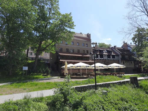 A picturesque outdoor café area with tables and large sun umbrellas situated in front of a series of old-style buildings. A well-maintained grassy area and trees surround the café, offering a serene and inviting atmosphere. The buildings have traditional architectural features with tiled roofs and wooden beams.