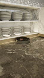 A robotic vacuum cleaner is positioned on a marble countertop beneath neatly stacked dishes on a shelf. The shelves have rows of plates and bowls, with an array of upside-down wine glasses on the top shelf. The marble has a mix of grey tones creating an elegant setting.