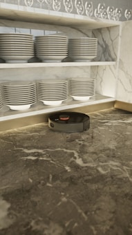 A robotic vacuum cleaner is positioned on a marble countertop beneath neatly stacked dishes on a shelf. The shelves have rows of plates and bowls, with an array of upside-down wine glasses on the top shelf. The marble has a mix of grey tones creating an elegant setting.