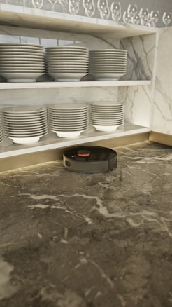 A robotic vacuum cleaner is positioned on a marble countertop beneath neatly stacked dishes on a shelf. The shelves have rows of plates and bowls, with an array of upside-down wine glasses on the top shelf. The marble has a mix of grey tones creating an elegant setting.