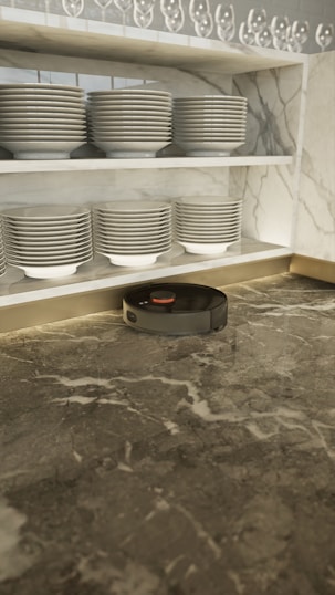 A robotic vacuum cleaner is positioned on a marble countertop beneath neatly stacked dishes on a shelf. The shelves have rows of plates and bowls, with an array of upside-down wine glasses on the top shelf. The marble has a mix of grey tones creating an elegant setting.