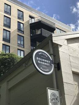 A modern building with a cream-colored facade and several large windows, alongside a black circular sign that reads 'Artisan Gourmet Gannet Bakers'. A smaller sign below with the word 'Supreme' is seen. The sky is clear with a few clouds, and some greenery is visible.