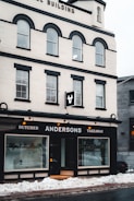 A multi-story building with a white facade and black accents houses a business named Andersons, which offers butcher and takeaway services. Large display windows show some indoor plants and the entrance is centrally located. Snow is piled up on the sidewalk in front of the building.