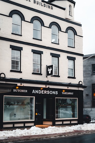 A multi-story building with a white facade and black accents houses a business named Andersons, which offers butcher and takeaway services. Large display windows show some indoor plants and the entrance is centrally located. Snow is piled up on the sidewalk in front of the building.