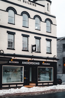 A multi-story building with a white facade and black accents houses a business named Andersons, which offers butcher and takeaway services. Large display windows show some indoor plants and the entrance is centrally located. Snow is piled up on the sidewalk in front of the building.