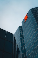Tall glass buildings with reflective windows are angled towards each other under an overcast sky. A bright red logo is prominently displayed on one of the buildings.