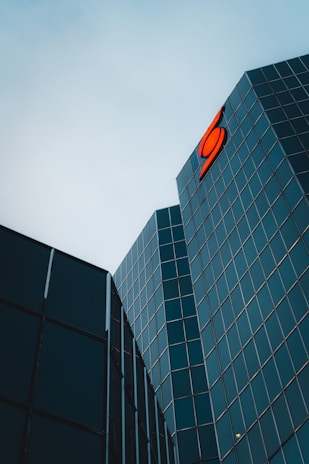 Tall glass buildings with reflective windows are angled towards each other under an overcast sky. A bright red logo is prominently displayed on one of the buildings.