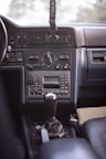 A view of a car's interior focusing on the central console and dashboard. The dashboard features several air vents, climate control knobs, and various buttons for controlling different functions. A gear shift is prominently placed in the foreground, and there is a partially visible lanyard hanging from above.