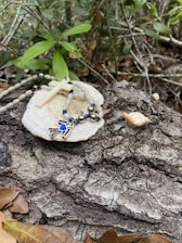 A rosary with a white cross and beads rests on a white stone-like surface placed on a textured tree bark. Green leaves and branches surround the scene, giving a natural and serene ambiance. Fallen brown leaves are scattered around, enhancing the rustic feel.