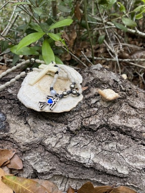 A rosary with a white cross and beads rests on a white stone-like surface placed on a textured tree bark. Green leaves and branches surround the scene, giving a natural and serene ambiance. Fallen brown leaves are scattered around, enhancing the rustic feel.